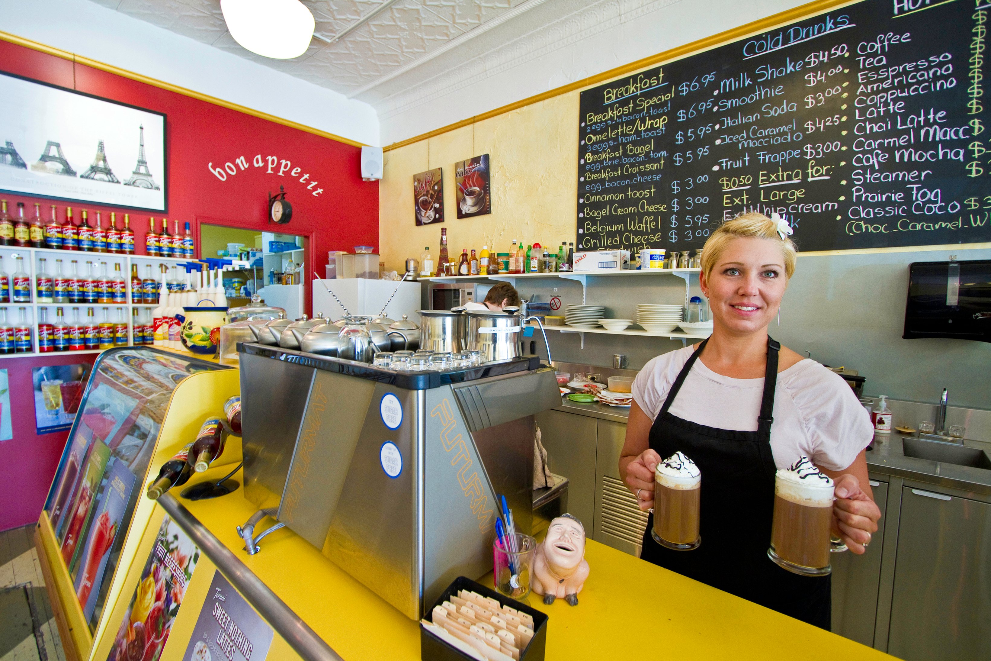 Blonde woman in white top and wearing black apron is serving two cafe lattes at a yellow counter with a commercial espresso machine on it and with a coffee menu on a blackboard on the wall behind.