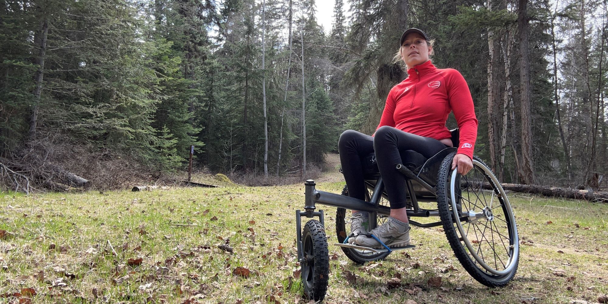 Lisa Franks, Paralympic athlete, woman with red shirt and black slacks sitting in a sports wheelchair capable of off-path travel, on a grass meadow with coniferous forest behind on an overcast day