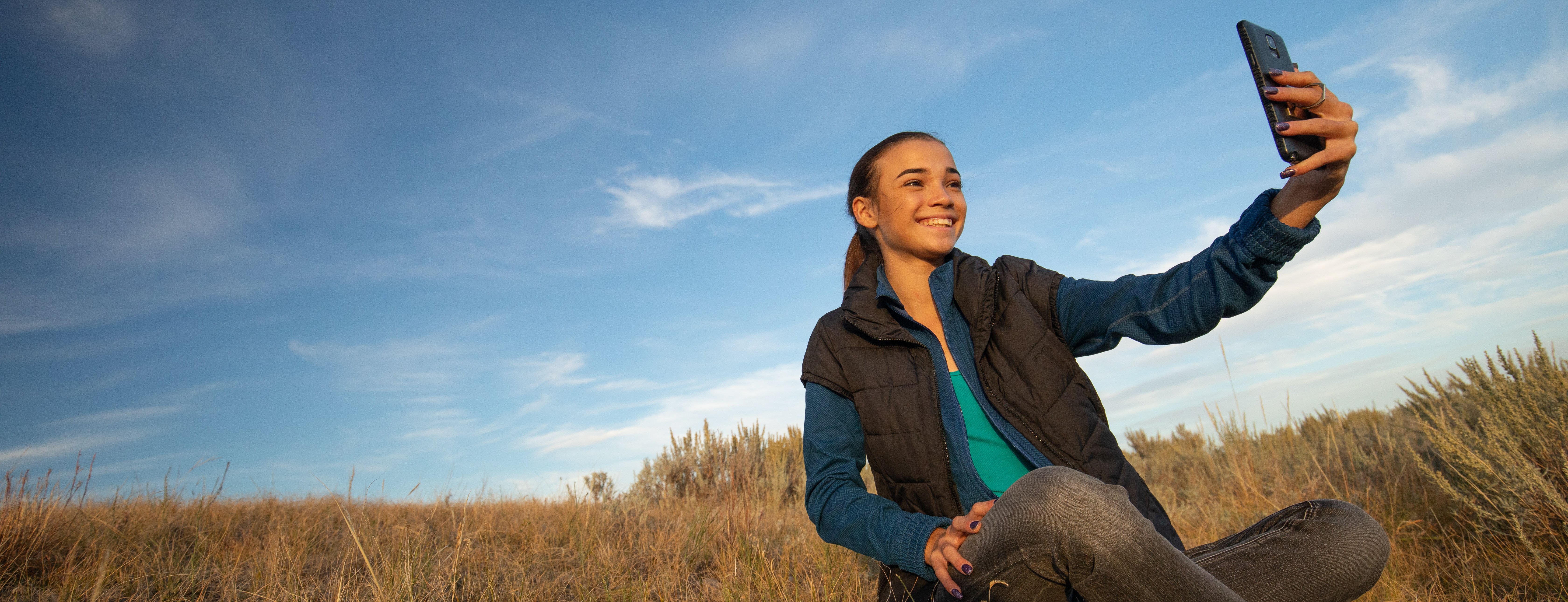 Smiling young woman lit by warm evening sunlight taking a phone selfie with natural grassland and bright blue sky behind