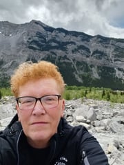 Alixe Rowe in dark coloured jacket standing at roadside pullout on Crowsnest Higway with Turtle Mountain and Frank Slide in background