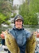 Carter Dueck sitting in a boat holding two fish against a backdrop of a northern boreal forest, raging river and frothy waterfall