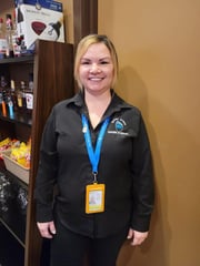 Charlene Standingready, wearing official name tage and dark shirt with "bear claw casino & hotel" logo embroidered near collar, standing next to a shelf with products sold at the business