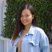 Thao Pham smiling at the camera while standing outdoors in a light blue jacket in front of a white fence and dark foliage