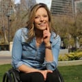 Lisa Franks, Paralympic athlete, smiling woman touching her chin with her left hand, wearing a blue shirt and black slacks, sitting in a wheelchair on grass in Victoria Park in downtown Regina with Hill Centre Towers in background on a sunny day