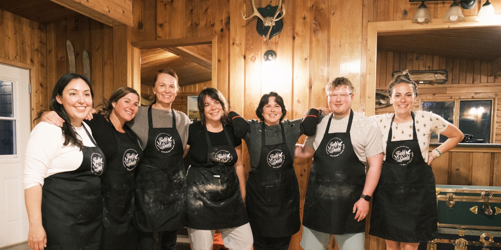 Seven people smiling broadly while standing next to each other with their arms interlocked over each others' shoulders in a room that has wood floor and wood panel walls decorated with a set of deer antlers and antique wooden cross-country skis