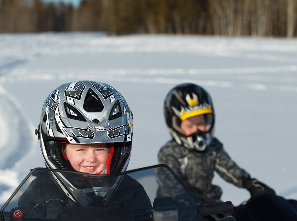 Two children on snowmobiles smiling at the camera.