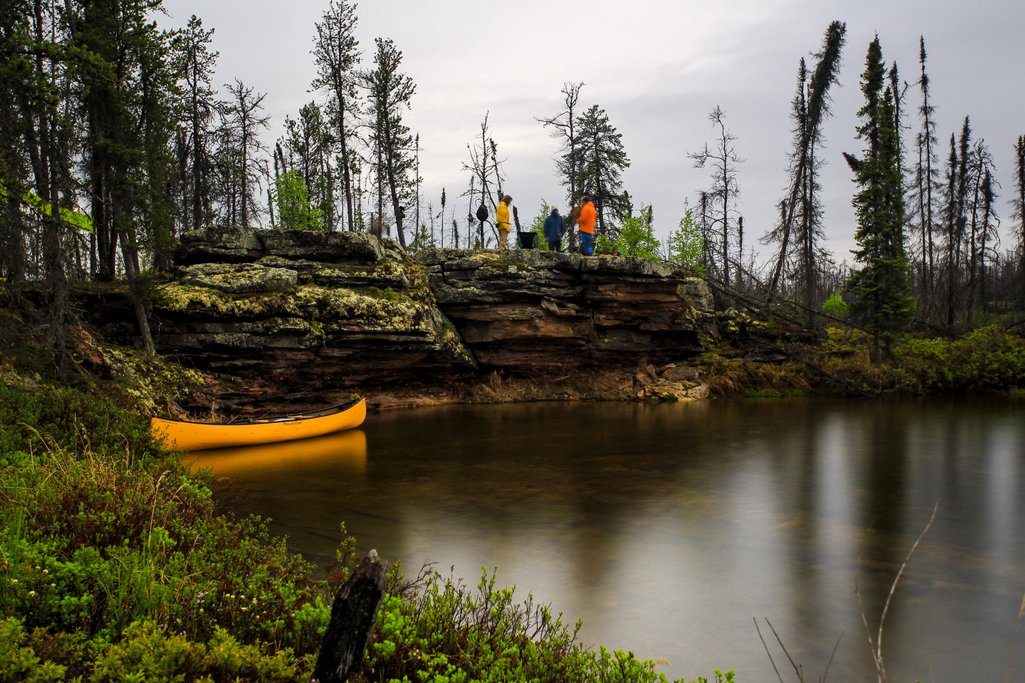 long exposure photo of a small cliff along a river with people standing on top and their canoe below.