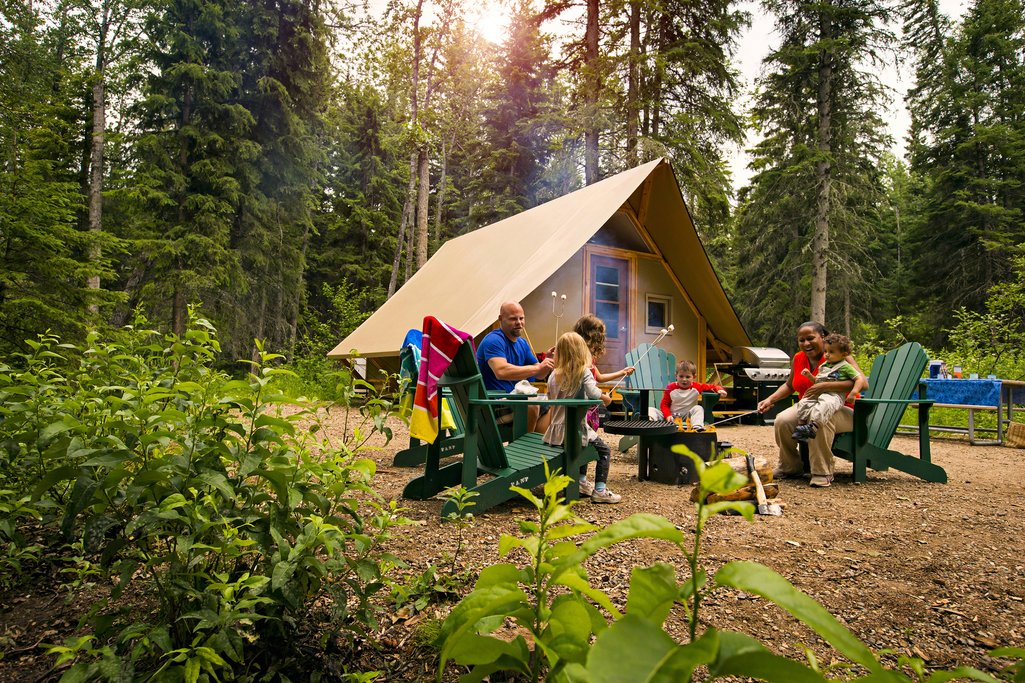 family gathered around a fire pit in the forest at a Parks Canada oTENTik campsite in Prince Albert National Park