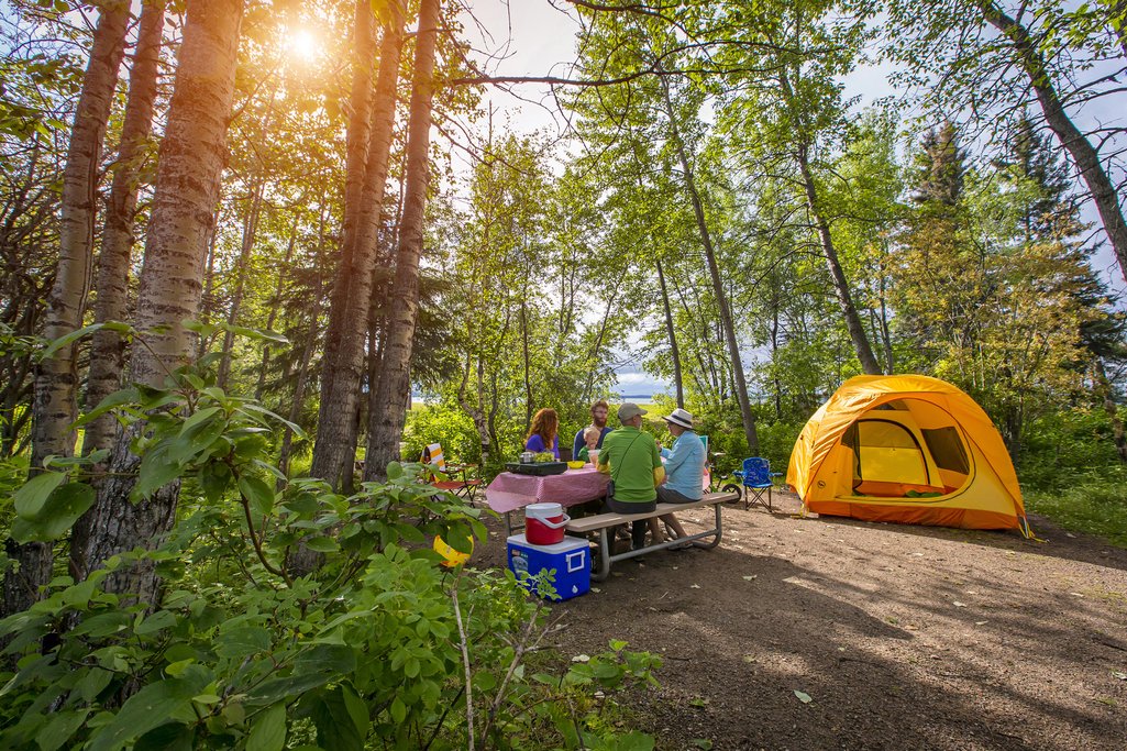 family sitting at a picnic table in their campsite beside their yellow tent surrounded by forest