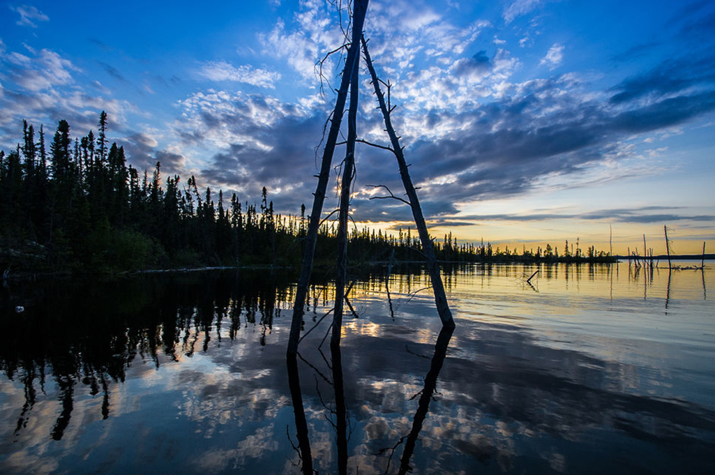 The sun sets on a beautiful evening in Tazin Lake, Saskatchewan. A structure constructed with three logs resembles a tipi sits in the water.
