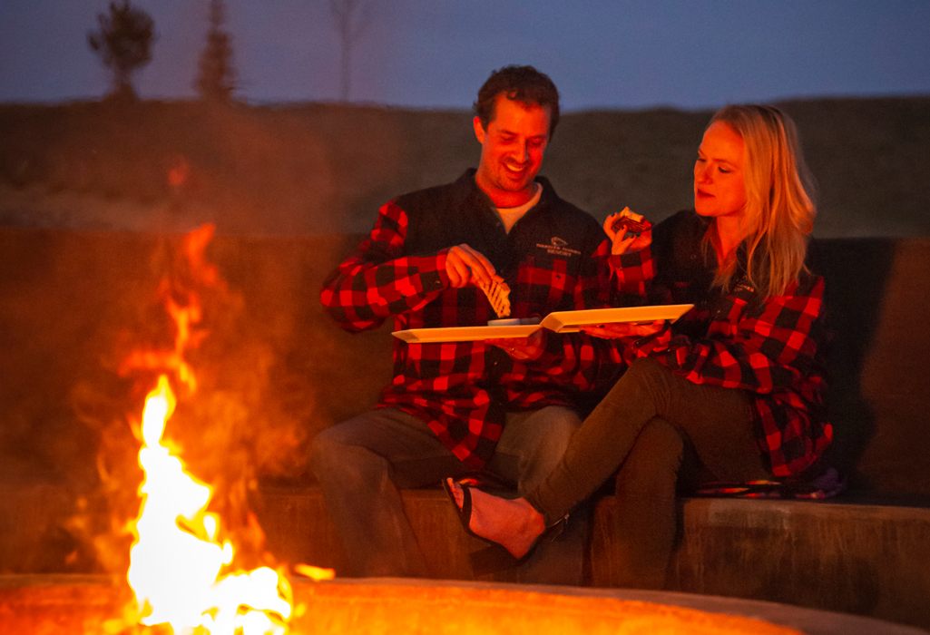 A couple in matching red plaid jackets enjoy s’mores by the outdoor firepit at Dakota Dunes Resort at dusk.
