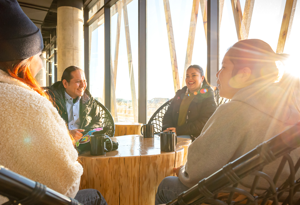 Four friends share coffee and conversation at Dakota Dunes Resort near Saskatoon, sitting by large sunlit windows overlooking the prairies.