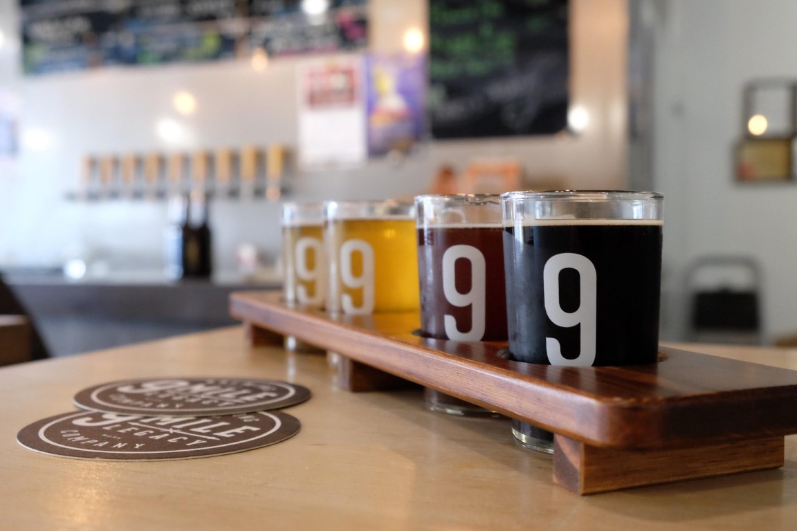 A flight of four craft beers on a wooden serving paddle next to brewing coasters in a brightly lit taproom