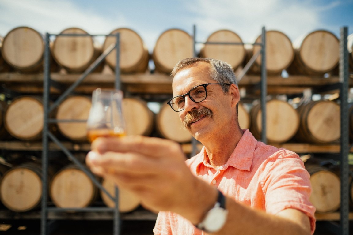 A distiller with glasses and a mustache inspecting a tasting glass of amber spirits outdoors in front of stacked wooden aging barrels
