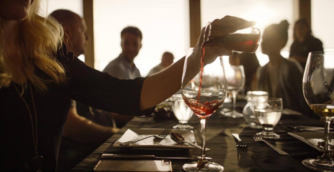 Close up of a hand pouring red wine from a small carafe into a glass at a sunset dinner event, with silhouettes of guests in the background