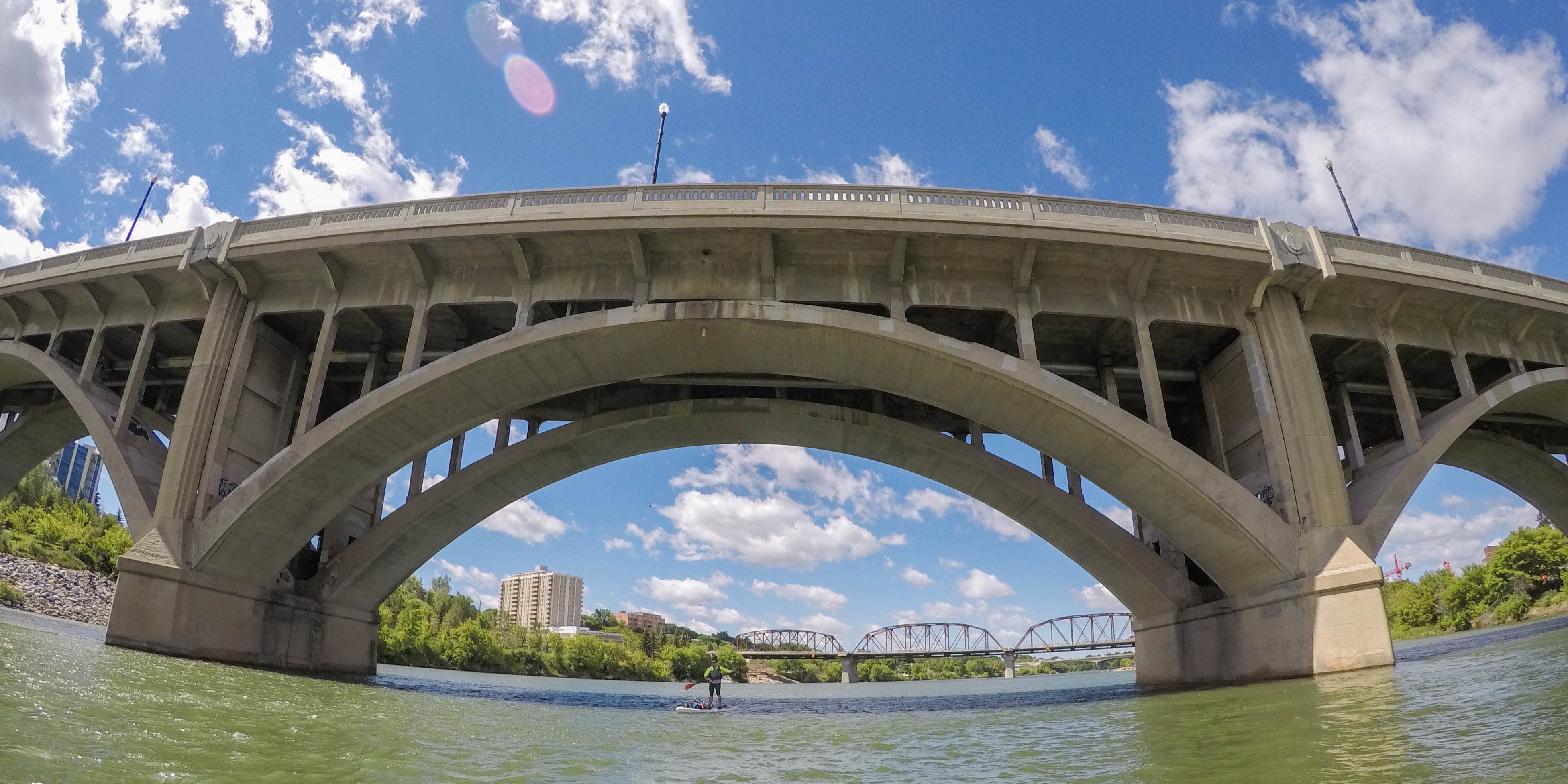 Broadway Bridge over South Saskatchewan River, Traffic Bridge and Nutana neighbourhood viewed from under the bridge arches on a bright sunny day with blue sky and scattered fluffy clouds