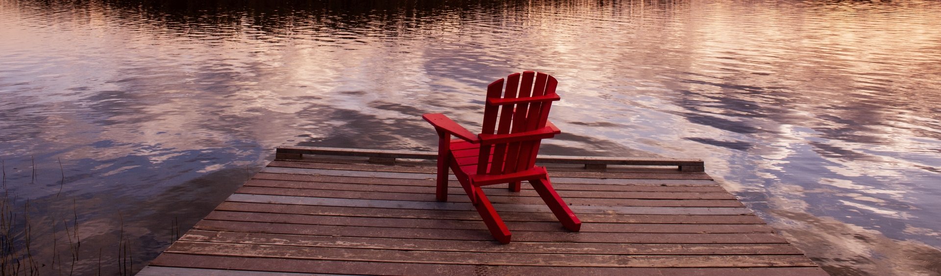 A red Adirondack chair on a dock with evening clouds reflected in the water.