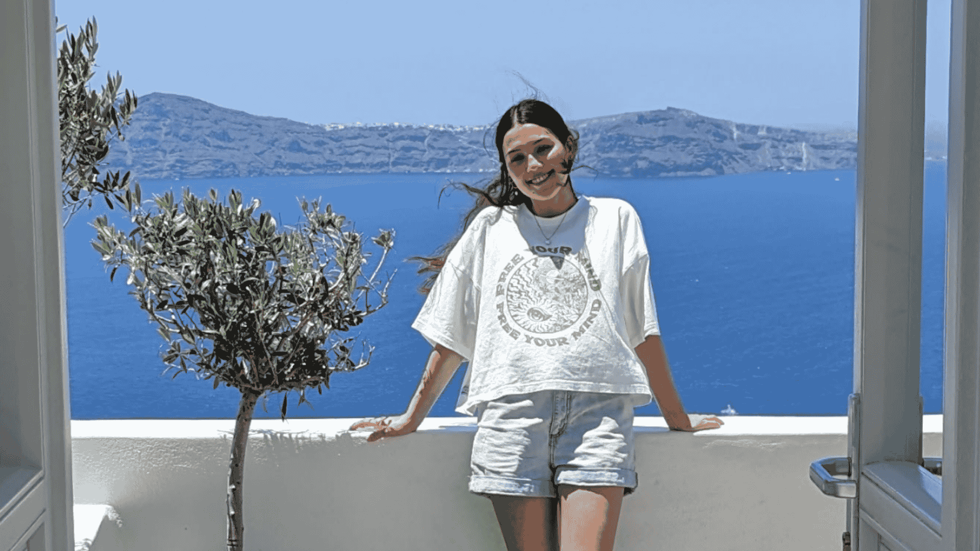 Traveller and content creator Gemma Edwards leans against a whitewashed Greek villa, overlooking a vividly azure Mediterranean ocean. In the foreground is a baby olive tree. Photo taken in Fira, Santorini, Greece
