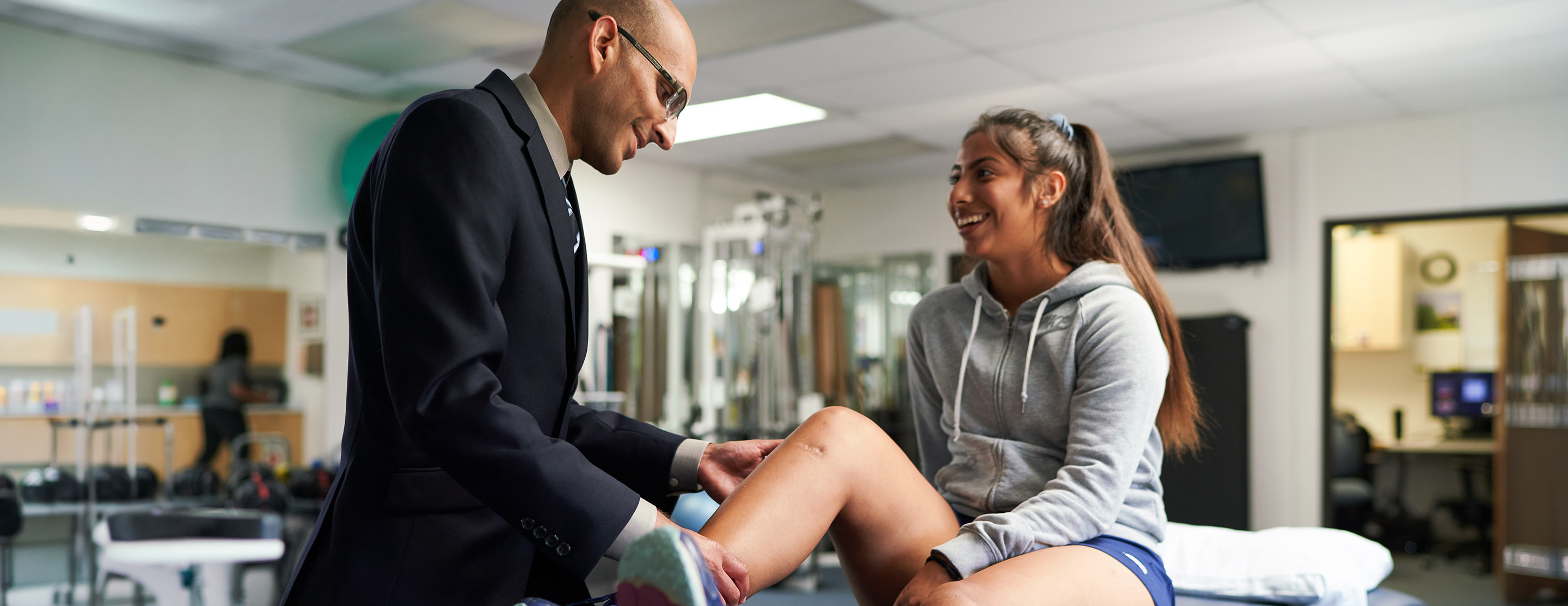 A clinician examines a smiling teenage athlete's knee as she sits on an exam table in a sports medicine clinic.
