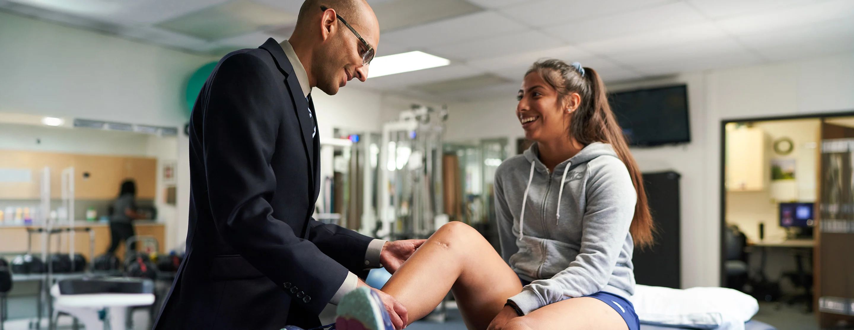 A clinician examines a smiling teenage athlete's knee as she sits on an exam table in a sports medicine clinic.