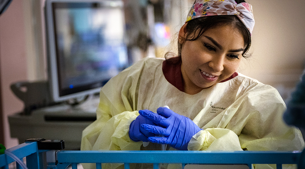 Roxana Gonzalez, in a protective gown and gloves, leans over an infant's bedside in the NICU.