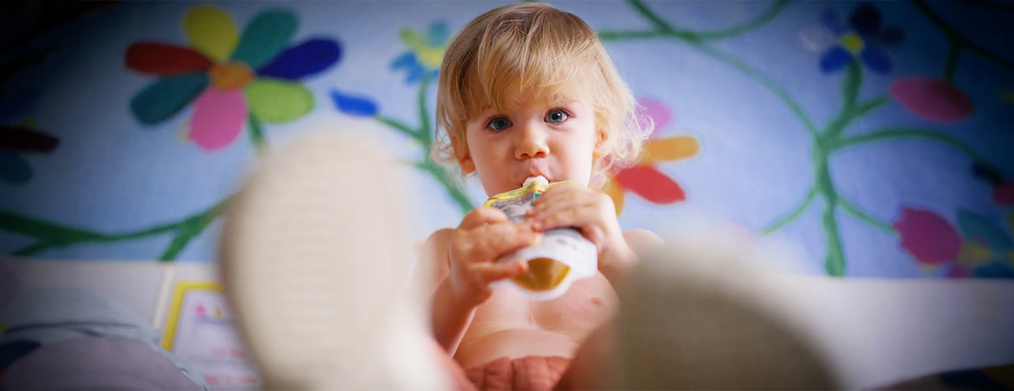 Miles, a toddler, sits on a colorful floral blanket drinking from a food pouch.