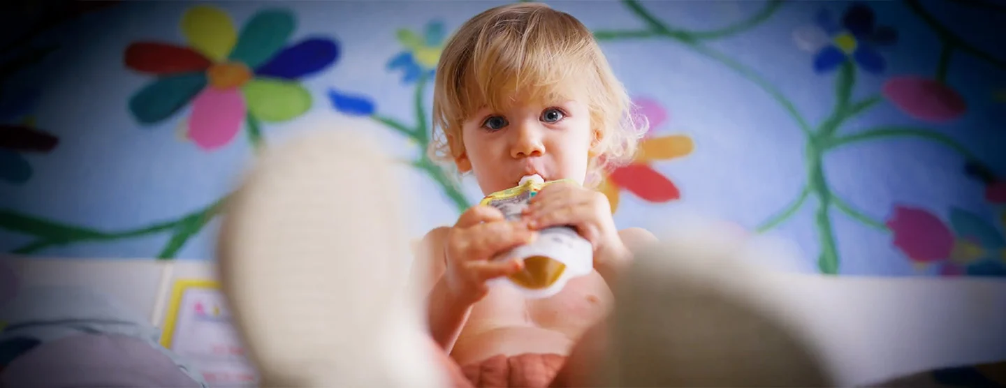 Miles, a toddler, sits on a colorful floral blanket drinking from a food pouch.