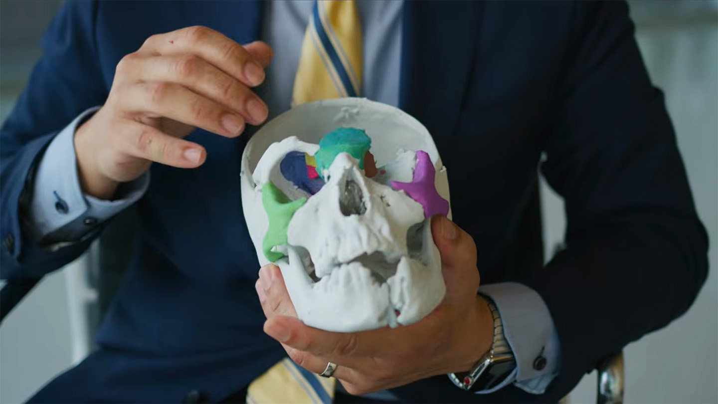 A surgeon holds a white 3D-printed model of a child's skull, with several facial bone segments printed in green, blue, teal and pink.
