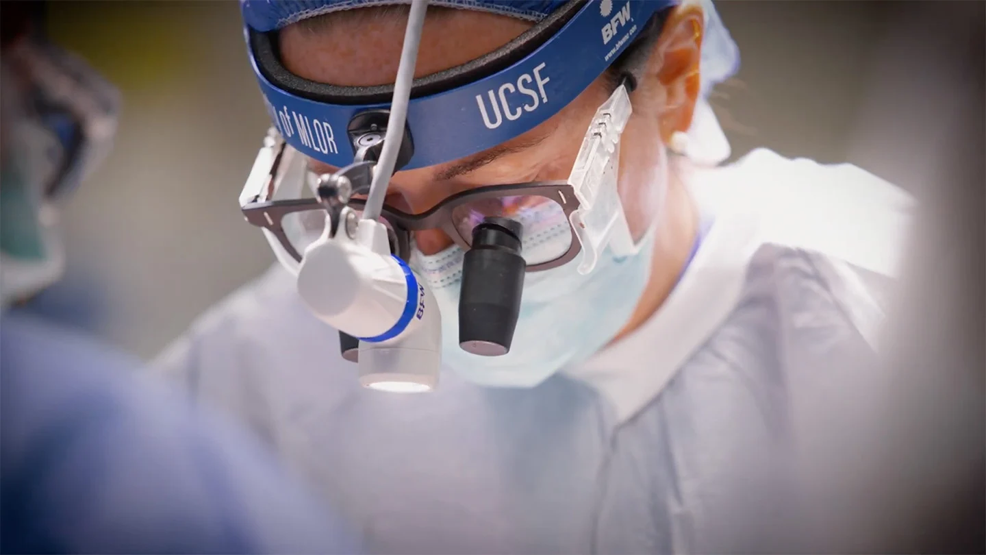 A surgeon wearing a UCSF headlamp and magnifying loupes looks down during an operation
