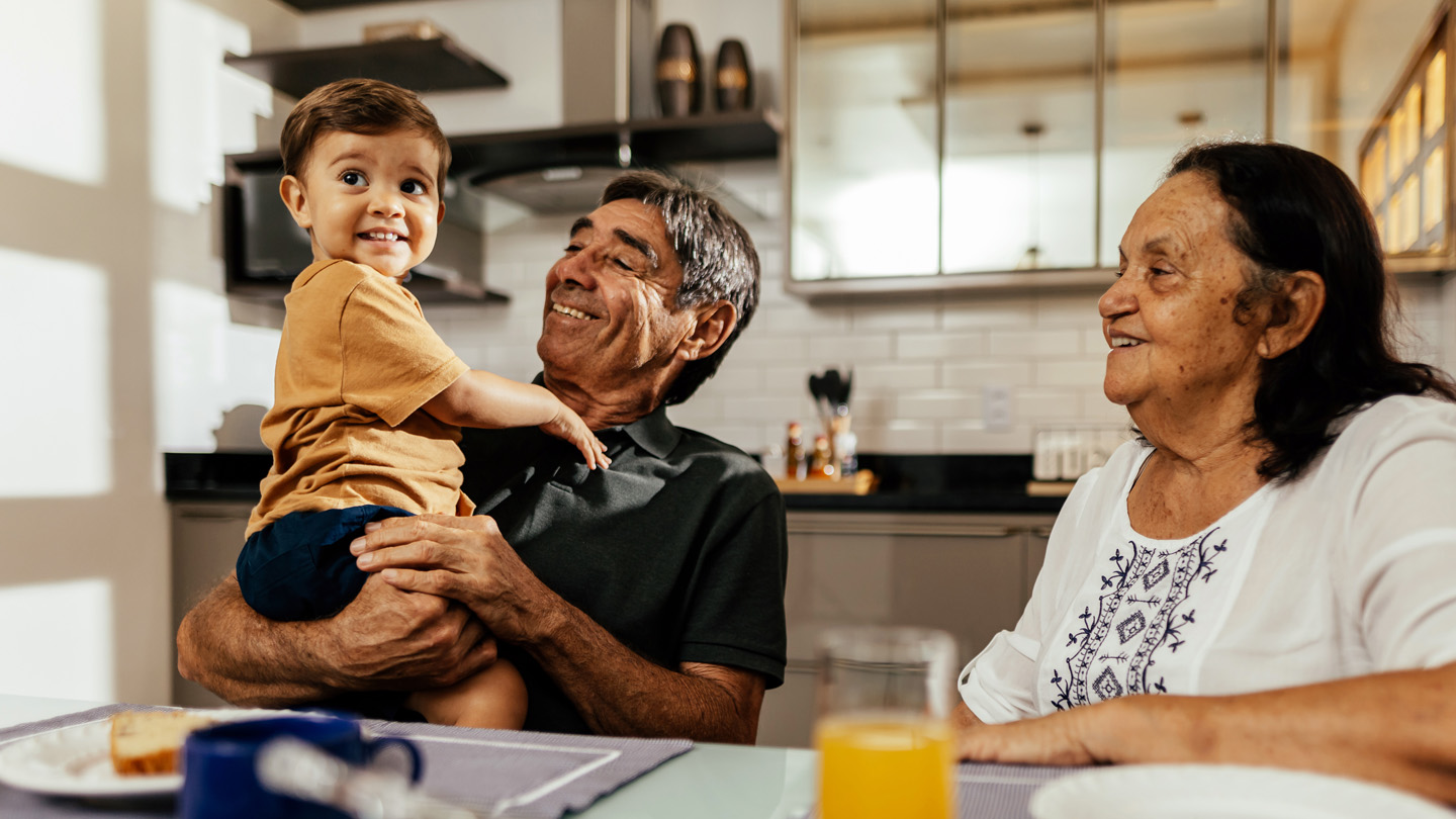 A grandfather holds a smiling toddler at a kitchen table while a grandmother sits beside them.