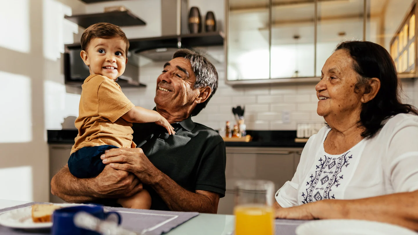 A grandfather holds a smiling toddler at a kitchen table while a grandmother sits beside them.