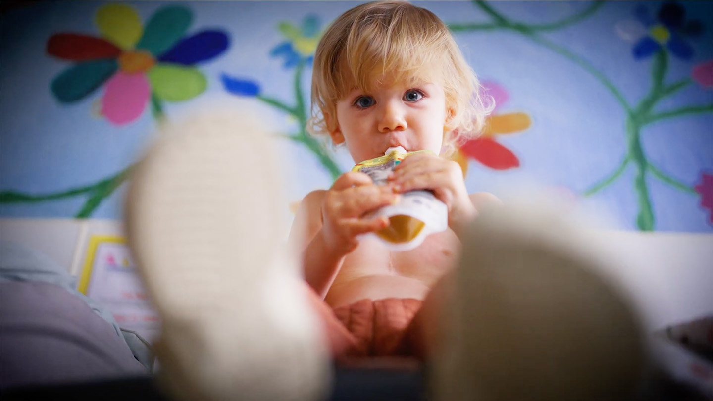 A toddler sips from a food pouch, sitting on a bed in front of a flower mural