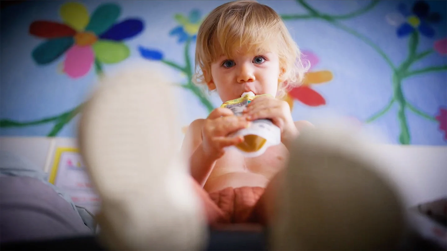 A toddler sips from a food pouch, sitting on a bed in front of a flower mural