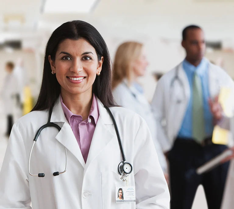 Smiling female doctor with dark hair and a stethoscope stands in a hospital, blurred colleagues in lab coats are in the background.