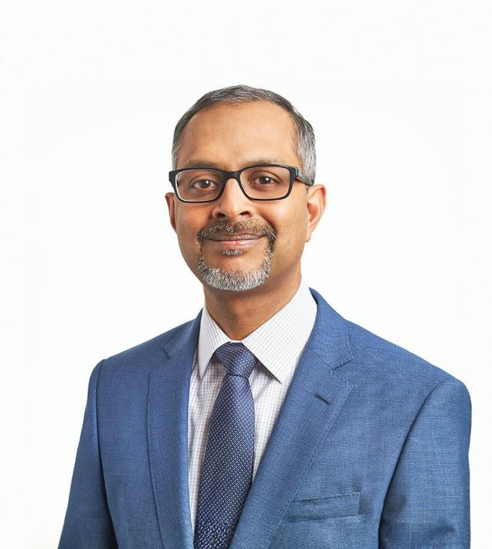 Portrait of a man with short gray hair and a beard, wearing glasses, a blue suit jacket, a white dress shirt, and a patterned tie, smiling slightly against a white background.