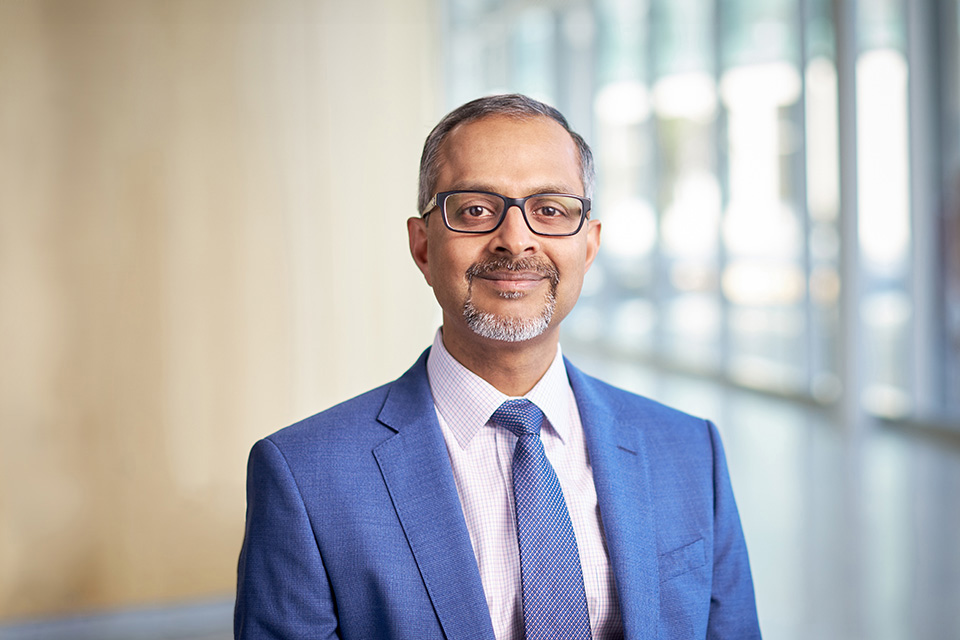 Portrait of a man with short gray hair and a beard, wearing glasses, a blue suit jacket, a white dress shirt, and a patterned tie, smiling slightly against a blurred background.
