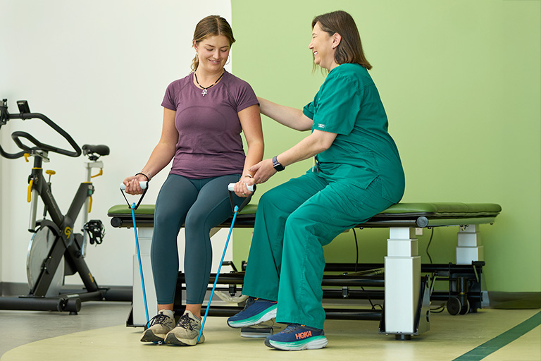 Image of a healthcare professional in a hospital setting, wearing a mask, apron, and cap, with medical equipment in the background.