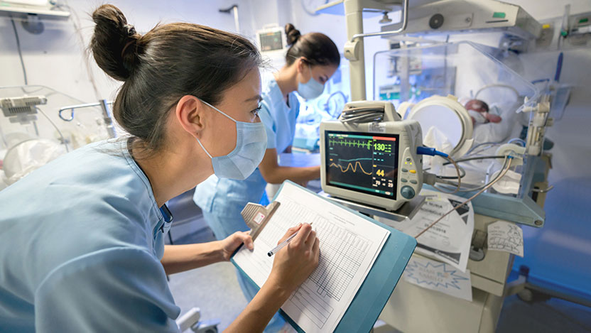 Nurse monitoring a premature newborn in an incubator while wearing a face mask