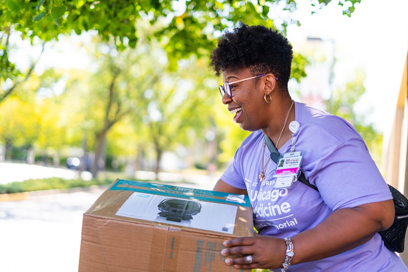 A smiling UChicago staff member in a purple shirt holds out a box containing an infant carseat