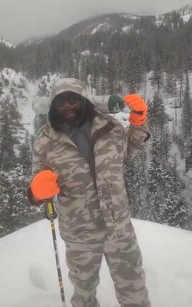 Leon Darby, a tall African-American man wearing a snowsuit and carrying an oxygen tank, stands smiling at the top of a snowy mountain with a view of treetops behind him.