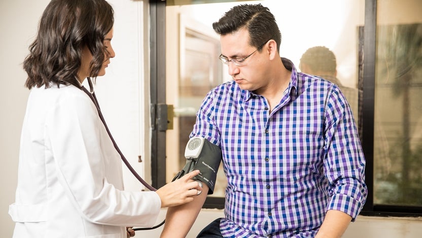 A female physician in a white coat uses a blood pressure cuff and stethoscope to measure the blood pressure of a man wearing business casual attire