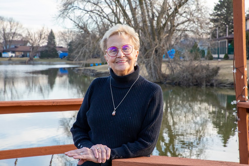 marcia pavich, an older white woman with short hair, stands smiling on front of a small lake or pond