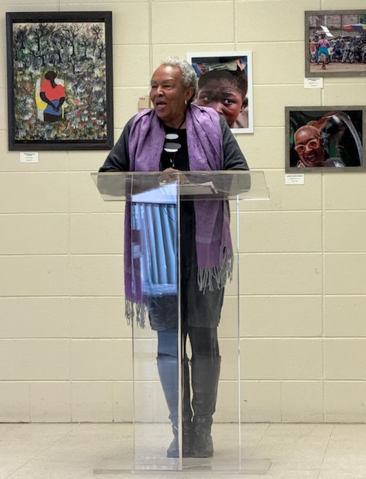 SuperAger Sheila Nicholes, 84, a Black woman with short silver hair, stands at a podium in front of some of her photography and speaks to attendees at her recent art showcase.