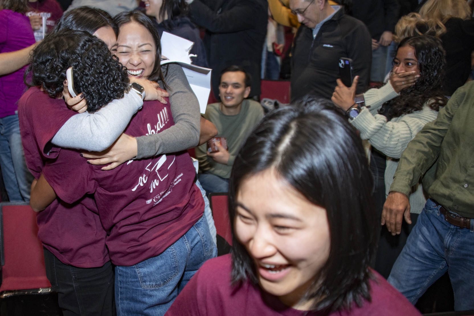 Pritzker med students in maroon shirts react to their match results. One girl grins in the foreground, while in the background three girls hug each other on one side and another holds up a cell phone on FaceTime while covering her mouth in a gesture of happy disbelief.