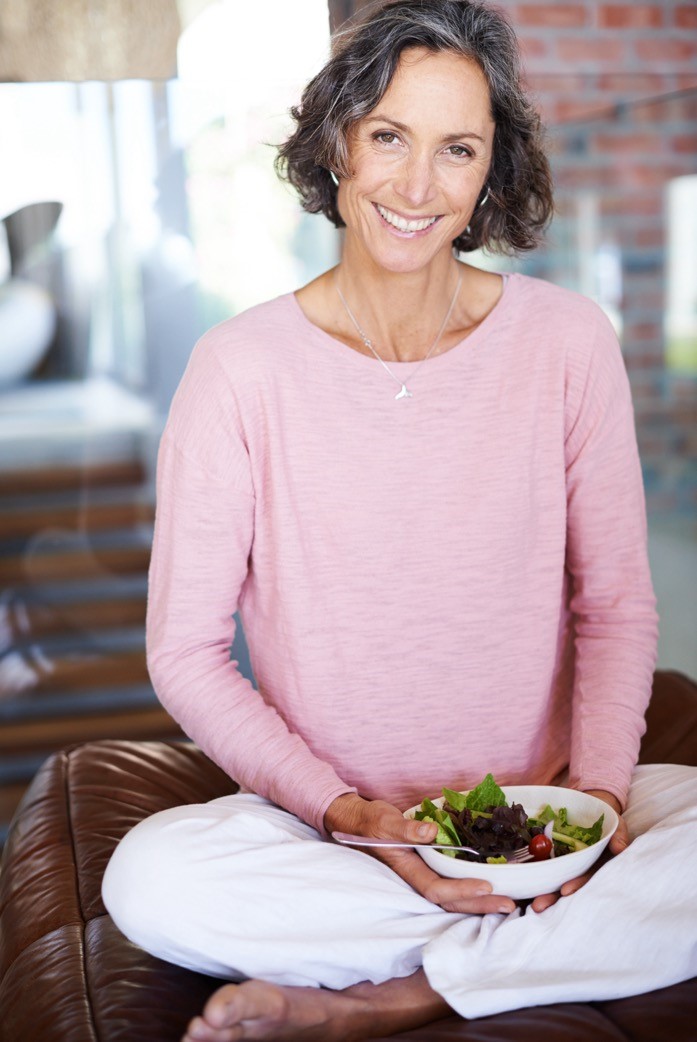 A woman in a pink shirt smiles while sitting with a bowl of salad in her lap.