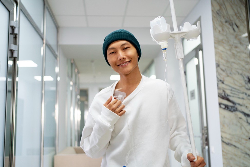 A young woman in a hospital gown standing with an IV drip smiling.