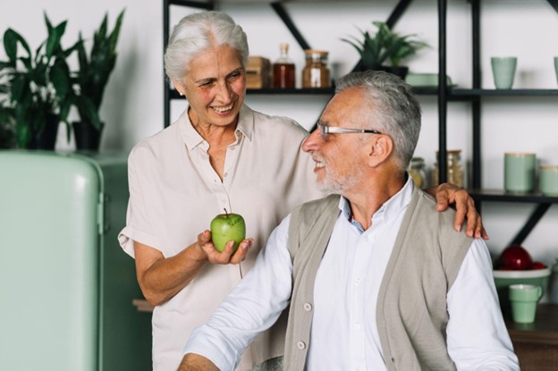 A happy elderly woman and man smiling together in a kitchen with a green apple representing healthy lifestyle habits