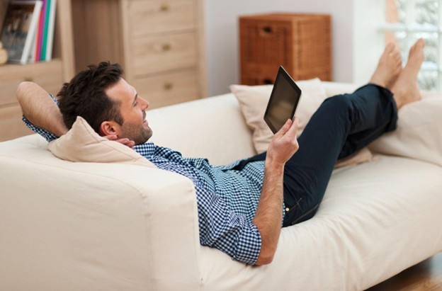 A man relaxing comfortably on a beige sofa while using a tablet, representing successful recovery and joint function