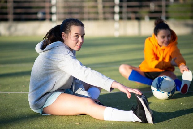 Two young female athletes stretching on a green sports field, highlighting the positive results of managing repetition and movement patterns.