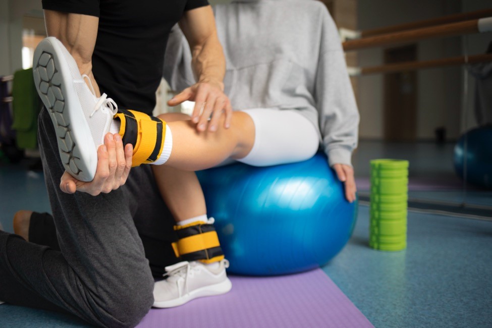 A woman sits on an exercise ball while a physical therapist helps her with knee rehabilitation exercises.